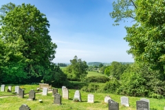 A rolling countryside view from the rear of the church. Gravestones and a bench can be viewed in the foreground.
